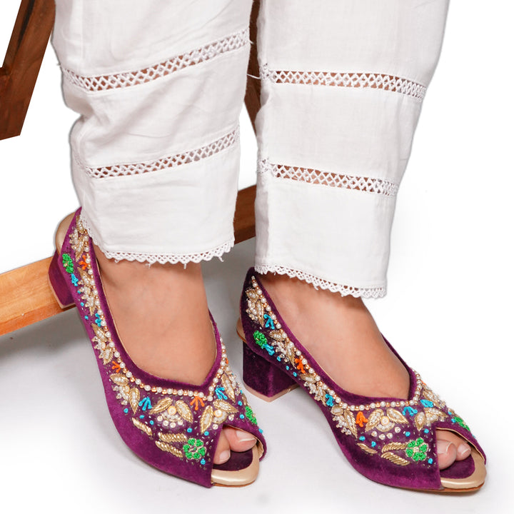 Purple embroidered shoes with gold and multicolor details worn with white pants.