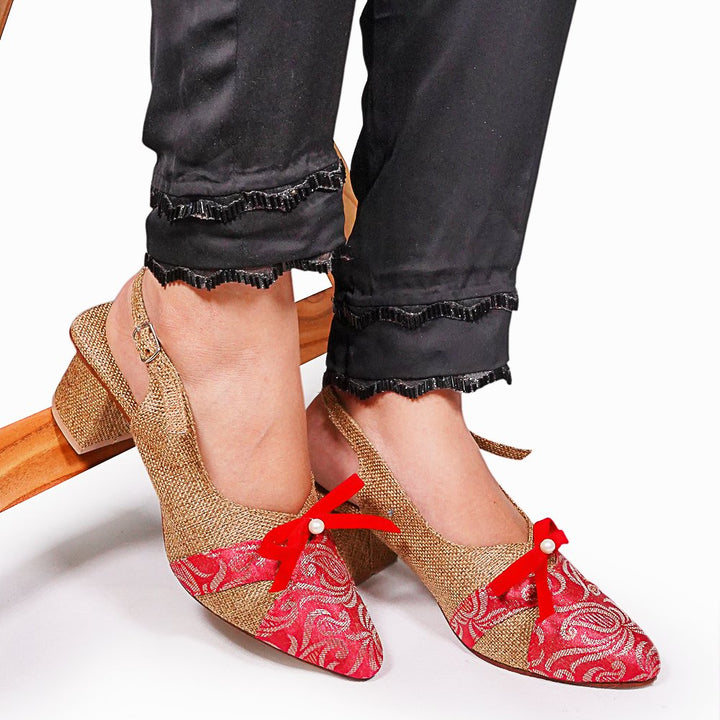 Red and gold patterned shoes with red bows worn by a person sitting on a wooden stool.