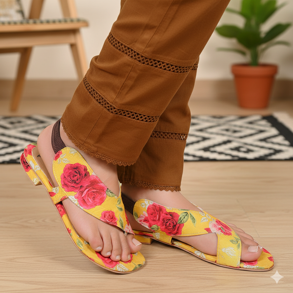 Person wearing yellow floral sandals with brown pants on a wooden floor.